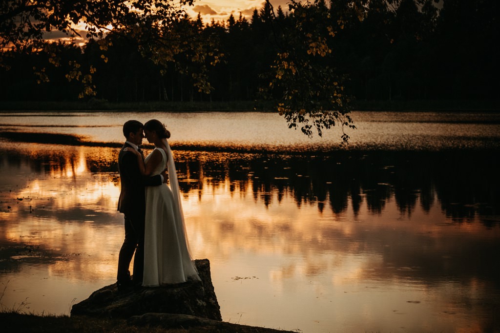 Ett brudpar som står på en setn i kanten av en sjö i solnedgången (under den gyllene timmen/golden hour).